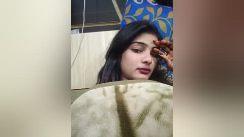 Media: Video of a young South Asian woman with dark hair, light skin, and makeup, wearing a blue floral dress. She is seen applying makeup in a reflective mirror, with a beige towel covering her face.