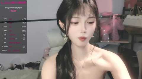 Media: Video of a young East Asian woman with pale skin and long black hair in a ponytail, wearing a strapless top, seated indoors. Background includes a desk with a pink bear, a laptop displaying streaming stats, and a messy room.