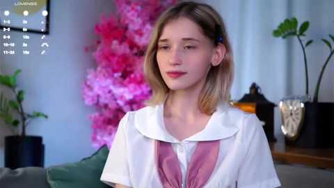 Media: Video of a young Caucasian woman with shoulder-length blonde hair, wearing a white sailor-style top with pink bow, in a softly lit room with pink floral backdrop, green plants, and a calendar.