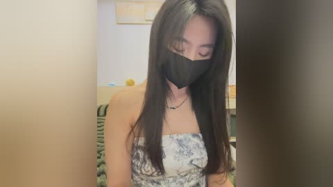 Media: A video of an Asian woman with long black hair, wearing a black face mask and a strapless floral dress, standing in a dimly lit room with a blurred background.