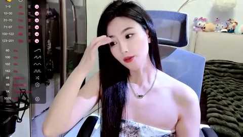 Media: Video of an Asian woman with long black hair and fair skin, wearing a strapless top, sitting on a blue chair, touching her face, in a bedroom with stuffed toys on a shelf.