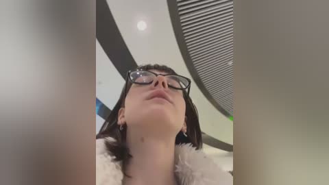 Media: Video of a woman with short brown hair, wearing black-framed glasses and a white fur coat, standing indoors with a modern, curved ceiling and a green light.