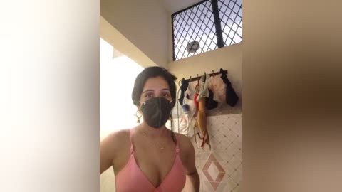 Media: A video of a woman with dark hair, wearing a black mask and pink bra, standing in a tiled bathroom with a window and clothes hanging on a rack.