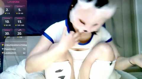 Media: Video of a young, light-skinned Asian woman in a white crop top and shorts, sitting on a bed, with blurred background. She has a small, round face and dark hair.