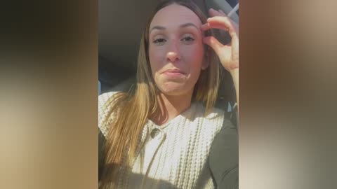 Media: Video of a young Caucasian woman with straight, light brown hair, wearing a white, textured sweater, sitting in a car. She has a serious expression, with sunlight creating a soft, natural lighting effect.