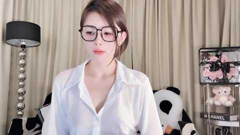 Media: A video of an Asian woman with fair skin and a slim physique, wearing black-framed glasses and a semi-transparent white blouse, standing in a room with beige curtains, a black lamp, and a Chanel perfume display.