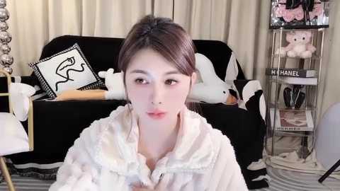 Media: Video of a young Asian woman with fair skin, brown hair, and a nose piercing, wearing a white fluffy robe, sitting in a chic, cluttered room with black furniture, white walls, and a Chanel perfume display.