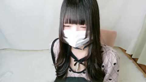 Media: A video of a young Asian woman with long black hair, wearing a white face mask, black choker, and a patterned blouse. She's seated indoors, against a plain white background.