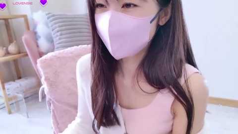 Media: A video of an Asian woman with long brown hair, wearing a pink mask and light pink top, sitting in a soft pink chair in a cozy, minimalist room with light-colored walls and wooden furniture.