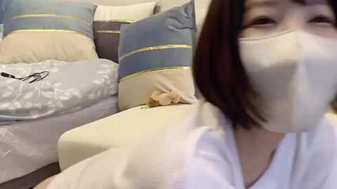 Media: Video of an Asian woman with short brown hair, wearing a white mask, lying on a bed with beige and blue striped pillows, and a teddy bear beside her.