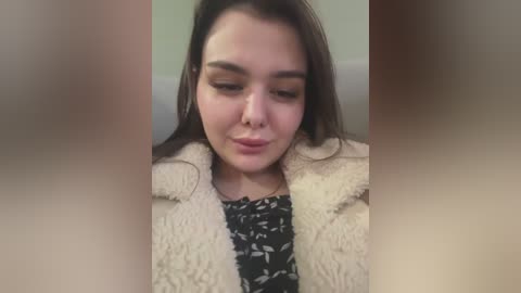 Media: Video of a young woman with long brown hair, wearing a black floral dress, under a beige fur coat. She has a nose ring and a neutral expression, indoors with blurred background.