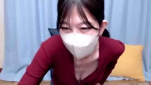 Media: Video of an East Asian woman with long black hair, wearing a white face mask, a red cardigan, and a black shirt, leaning forward, with light blue curtains in the background.