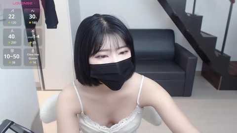 Media: Video of an East Asian woman with straight black hair, wearing a black face mask and a white lace-trimmed camisole, sitting indoors near a black couch and stairs.