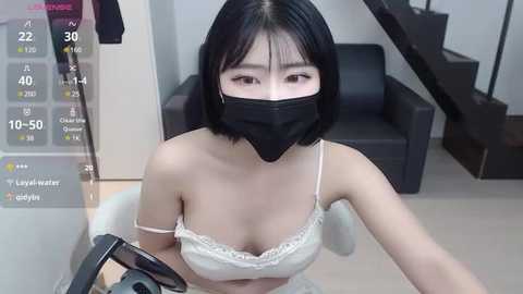 Media: A young East Asian woman with short black hair, fair skin, and a black face mask rides a motorcycle, wearing a white lace bra and beige pants in a modern, minimalist room with a black leather couch and a staircase.