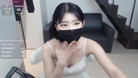 Media: Video of an East Asian woman with straight black hair, wearing a black face mask, white lace camisole, and sitting in a modern, minimalist living room with a black leather sofa and wooden staircase.