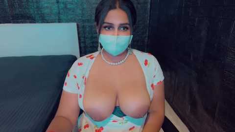 Media: Video of a plus-sized woman with large breasts, wearing a floral dress with deep cleavage, a face mask, and pearl necklace, in a dimly lit room with a brick wall.