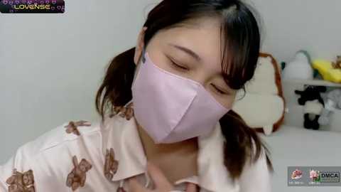 Media: A video of an Asian woman with straight black hair, wearing a light purple face mask and a floral-patterned blouse, sitting indoors with a blurred background featuring stuffed toys.