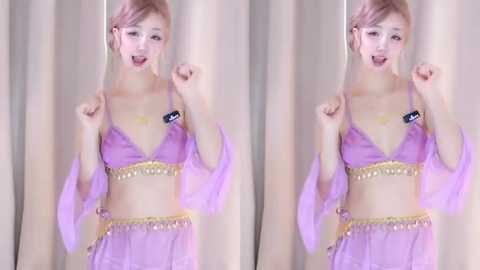 Media: Video of a smiling, slender Asian woman with fair skin and light pink hair, wearing a see-through, lavender bralette with gold trim and matching sheer skirt. She poses playfully with fists raised, against a beige curtain backdrop.