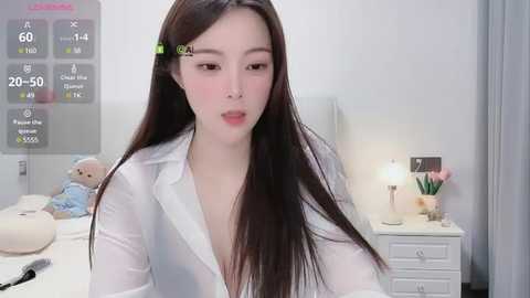 Media: Video of an Asian woman with long black hair, wearing a white blouse, in a modern bedroom with white walls, a teddy bear, and a digital clock displaying 20:50.