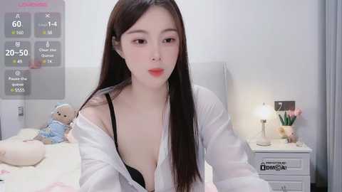 Media: A video of an Asian woman with long black hair, fair skin, and red lipstick, wearing a white lab coat and black bra, sitting on a bed with teddy bears, a nightstand with a lamp, and a digital clock.