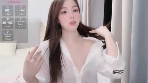 Media: A video of a young East Asian woman with long, straight brown hair, wearing a white, open-buttoned shirt, posing in a modern, minimalistic bedroom with grey curtains and a white bed.