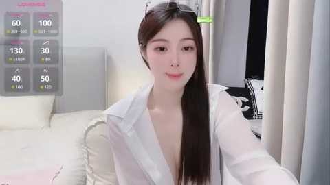 Media: Video of an East Asian woman with long, straight dark hair, fair skin, and a slim physique, wearing a see-through white shirt, sitting on a bed. The room features modern, minimalistic decor with white and gray elements.