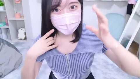 Media: Video of an Asian woman with long black hair and light skin, wearing a light blue striped top and a pink mask, gesturing with both hands, in a bright, modern bedroom with white furniture and a plush bed.