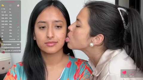 Media: Video of two women, one Asian with long black hair, wearing a colorful top, and the other with long black hair, kissing her cheek. Background shows a digital clock and a blurred room.