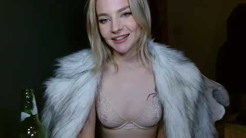 Media: Video of a smiling blonde woman in a white fur coat, revealing a lacy pink bra, against a dark background.