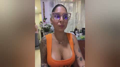 Media: A video of a tattooed woman with large breasts, wearing orange tank top, glasses, and dark hair tied back, in a cluttered, dimly lit room with floral decor and a window.