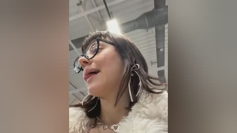 Media: Video of a young woman with glasses and a heart-shaped pendant, wearing a white fur-trimmed coat, against a modern industrial background with exposed pipes and ceiling lights.