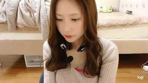 Media: Video of a young Asian woman with long brown hair, wearing headphones, seated on a wooden floor in a bedroom with a white bed, cream-colored pillows, and a stuffed animal.