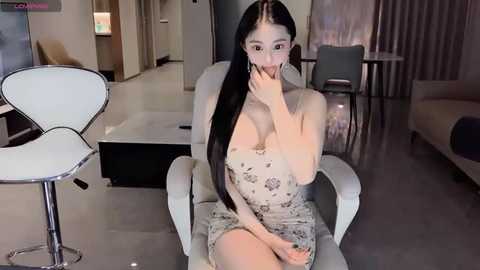 Media: Video of an East Asian woman with long black hair, slender physique, and fair skin, wearing a strapless floral dress, seated in a modern living room with white furniture and a minimalist decor.