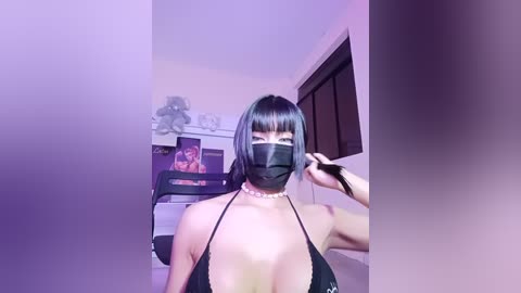 Media: Video of an Asian woman with straight black hair and bangs, wearing a black bikini top, black face mask, and pearl necklace, in a dimly lit bedroom with purple lights and a TV.