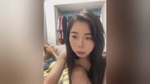 Media: A video of a young Asian woman with long black hair, wearing a pink top, taking a selfie in a bedroom with a bed and hanging clothes in the background.