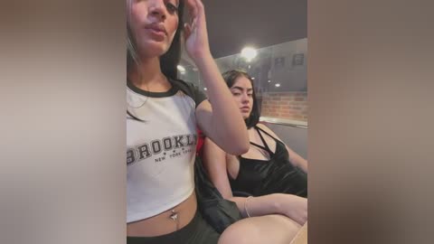 Media: Video of two women in a dimly lit bar. One woman in a white Brooklyn T-shirt with black trim and black shorts, the other in a black dress. Brick wall and ceiling lights in background.