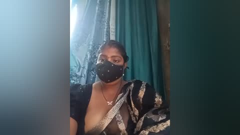 Media: A video of a South Asian woman in traditional attire, wearing a black mask, black sari with gold embroidery, and a necklace. Background features teal curtains and a window with greenery.