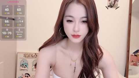 Media: Video of an East Asian woman with long, wavy brown hair, fair skin, wearing a white tank top and large hoop earrings, standing indoors.