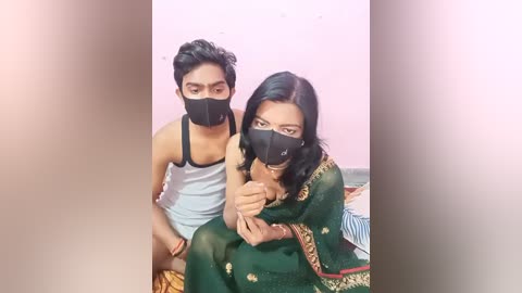 Media: A video of a young South Asian couple wearing masks, the man in a white tank top and shorts, the woman in a green sari with gold embroidery, sitting closely on a bed.