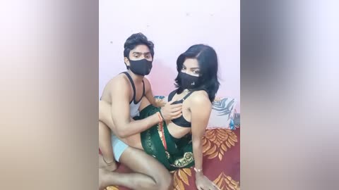 Media: Video of a man and woman in Indian traditional attire, wearing face masks, sitting closely on a red and yellow patterned bedspread.