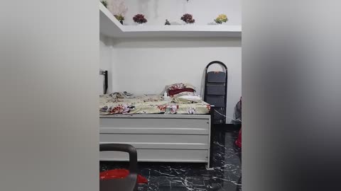 Media: Video of a small, sparsely furnished room with a white bed covered in floral bedding, a black marble floor, a white shelf with flowers, and a black standing fan.