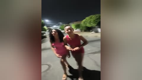 Media: Video of two young women with medium skin tone and long dark hair, dressed in matching pink swimsuits, standing outdoors at night. Background shows a residential street with a lit streetlamp, blurred bushes, and parked cars.