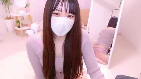 Media: Video of an East Asian woman with long brown hair, wearing a white mask, light gray cardigan, and white gloves, seated in a bright, minimalist room with white walls and a mirror.