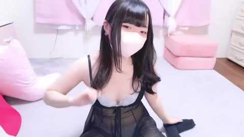 Media: Video of an East Asian woman with long black hair, fair skin, and wearing a black sheer lingerie set with a white lace bra, sitting on a light gray mat in a pink room with soft furnishings.