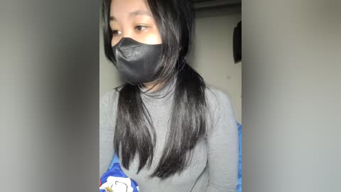 Media: Video of a young Asian woman with long black hair in pigtails, wearing a gray long-sleeve shirt, black face mask, and blue and white patterned gloves, indoors against a plain gray wall.