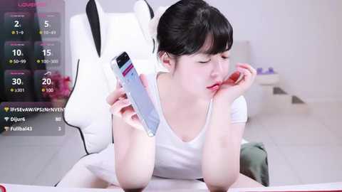 Media: A video of an Asian woman with black hair, wearing a white tank top, sitting on a white chair, holding a smartphone, in a minimalist room with a digital screen overlay.