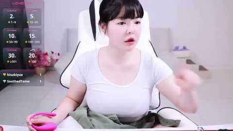 Media: Video of a young East Asian woman with black hair in a ponytail, wearing a white t-shirt, seated on a white gaming chair in a minimalistic, white room.