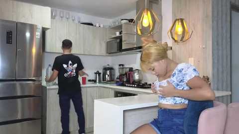 Media: Video of a modern kitchen with a blonde woman in blue denim shorts and a floral crop top cooking, a man in a black Adidas shirt and dark pants standing at the counter, and two pendant lights.