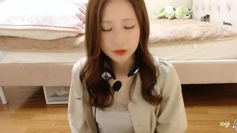 Media: A video of an Asian woman with long brown hair, wearing headphones and a beige cardigan, sitting on a wooden floor in a softly lit bedroom.