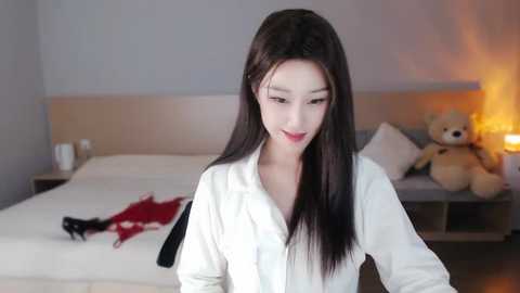 Media: A video of an East Asian woman with long, straight black hair, fair skin, and red lipstick, wearing a white button-up shirt, smiling in a modern bedroom with a beige bed, teddy bear, and a red garment on the floor.